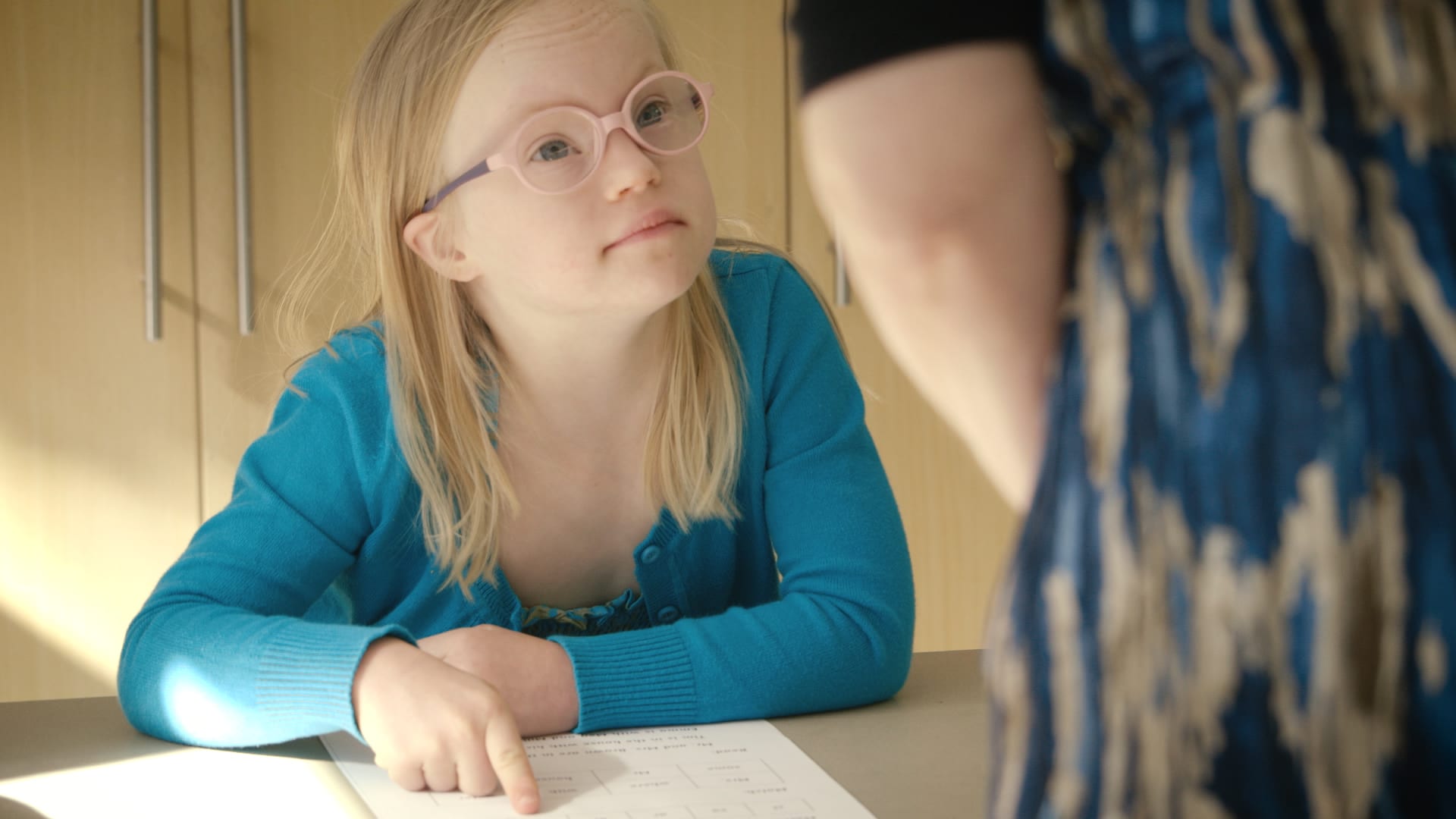 A young girl sits at a desk and a teacher helps her. The little girl has Down's syndrome
