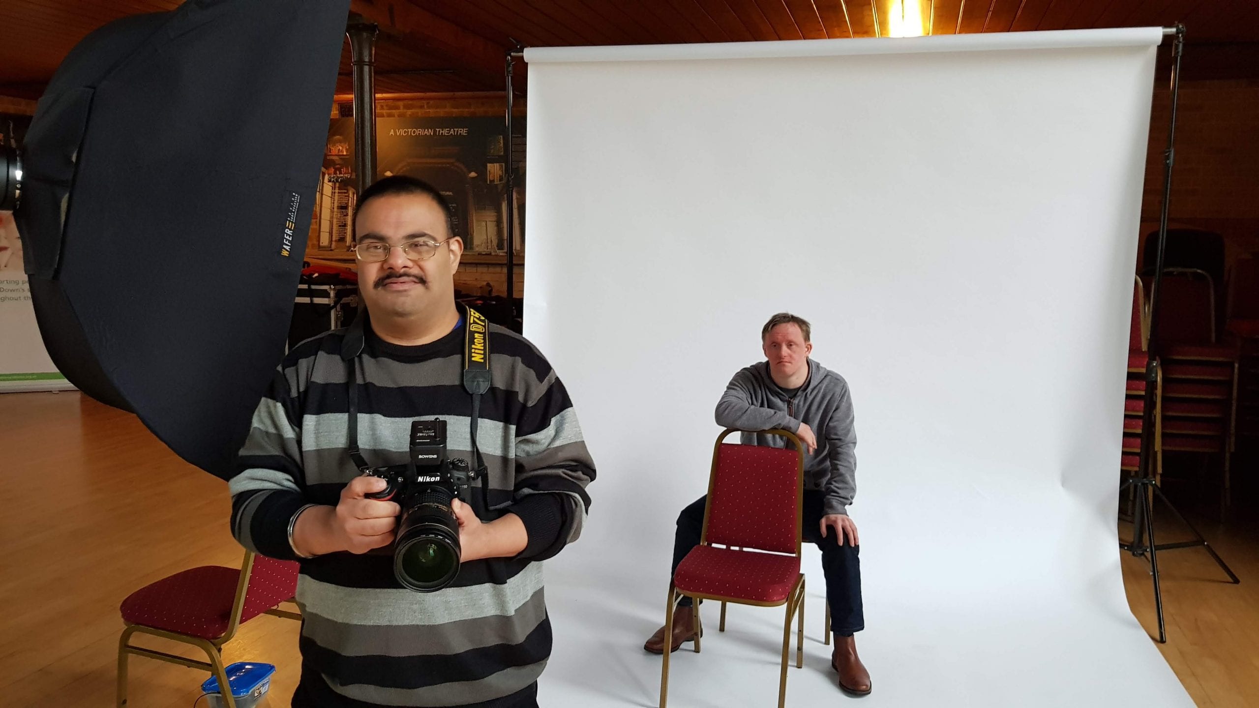 A man who has Down's syndrome poses to be photographed. He sits on a chair in front of a white backdrop. The photographer faces the cameraman also has Down's syndrome