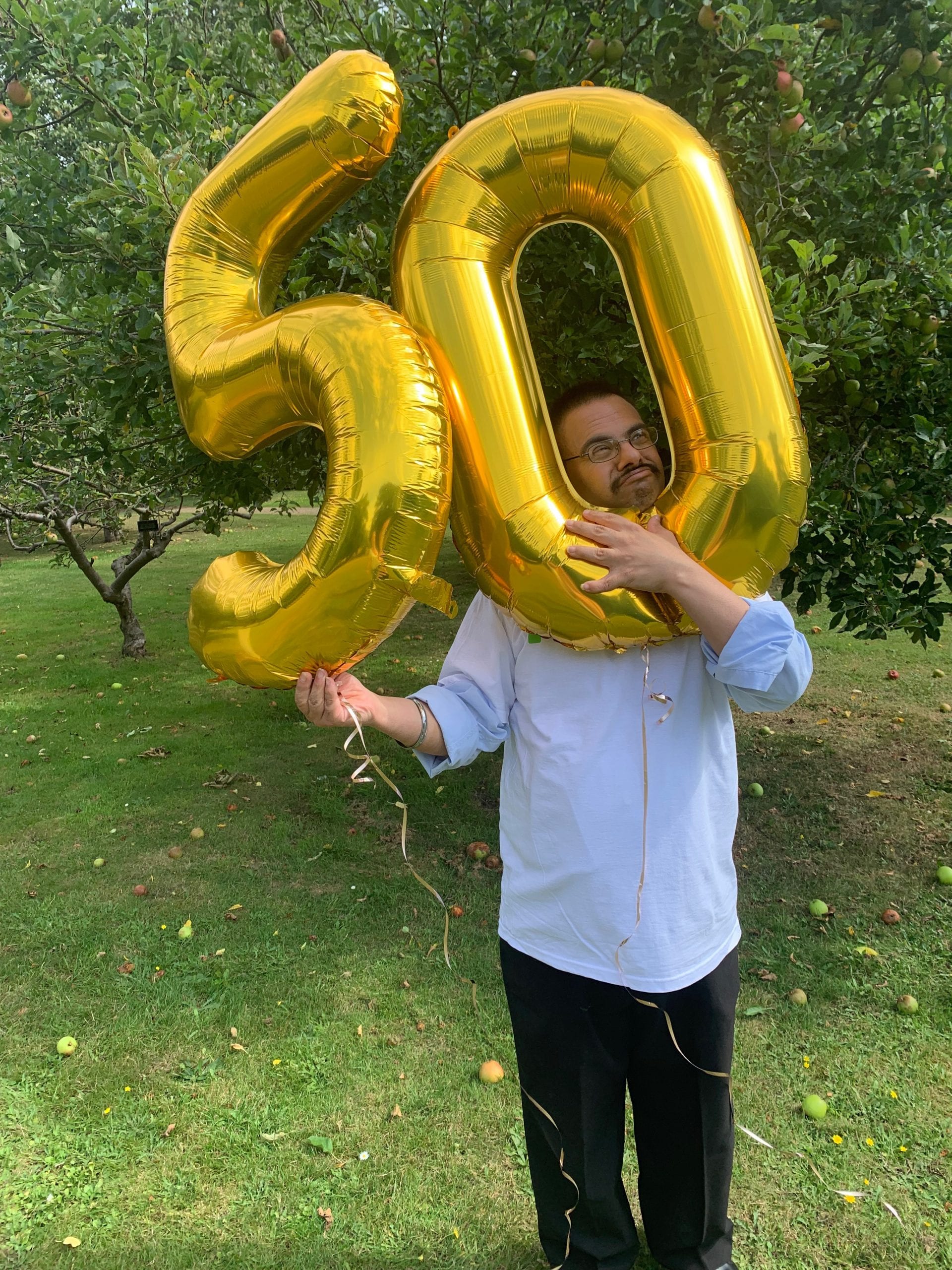 A man who has Down's syndrome stands in a garden holding two large, golden helium baloons that make up the number fifty.