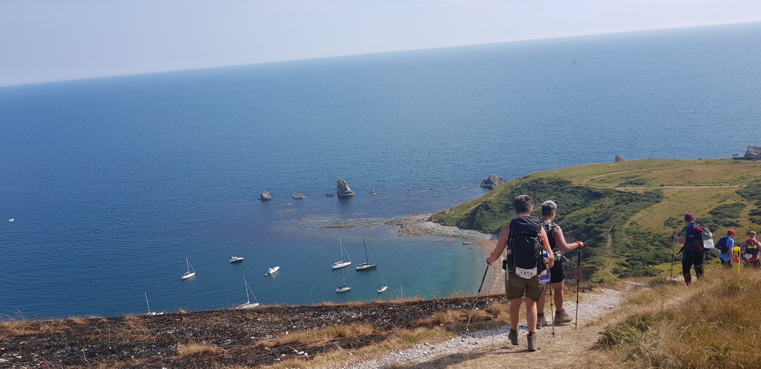 A group of walkers, using poles, walks down a steep coastal path. The sea is visible in the background. Its a sunny day and the sea is blue.