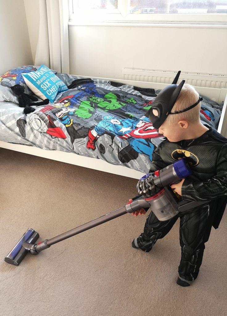 A young boy in a Batman outfit vacuums the carpet in his bedroom. His bed has a superhero duvet cover