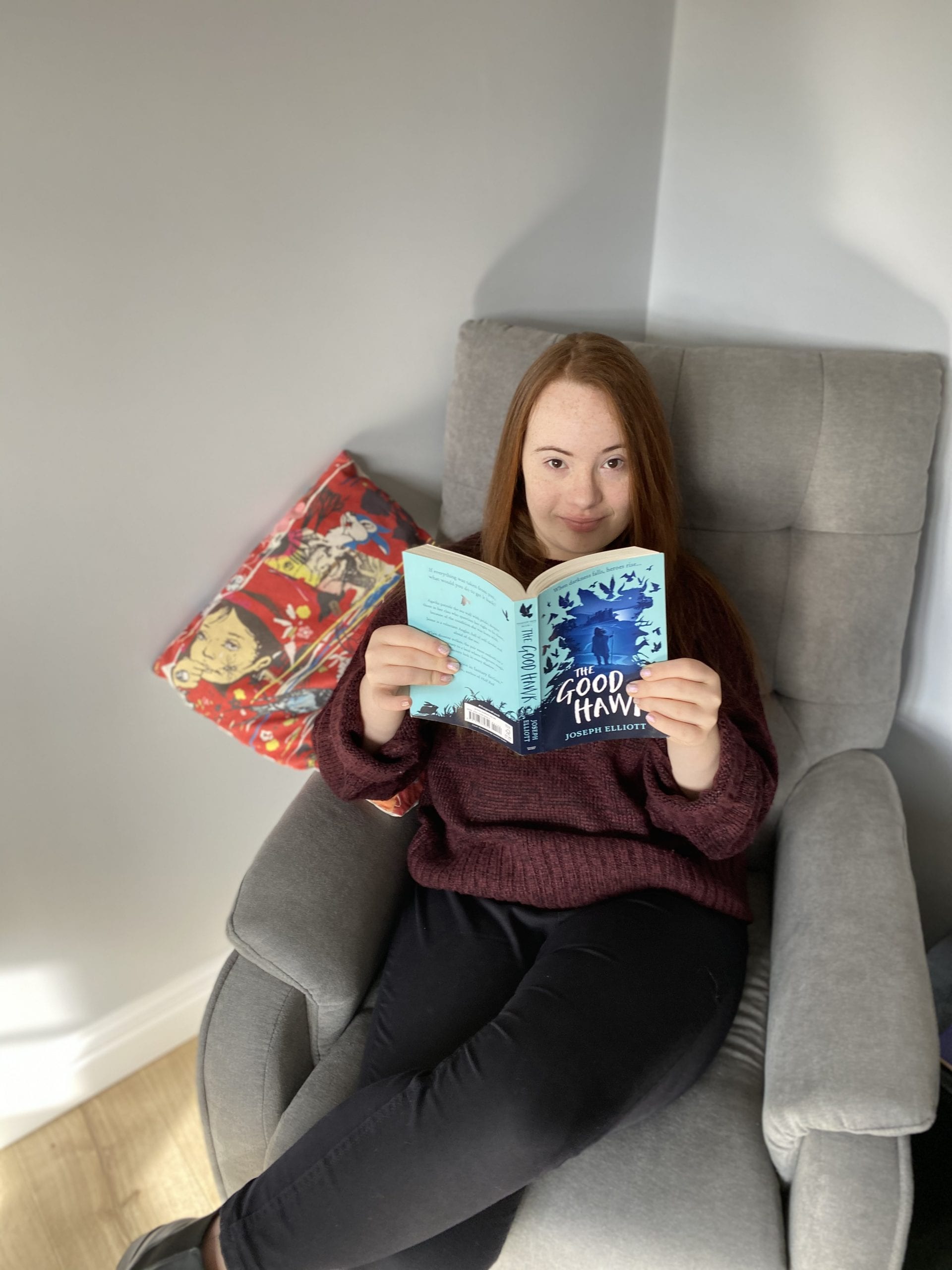 A woman sits in a grey easy chair holding an open paperback book. She is looking at the camera. The young woman has Down's syndrome