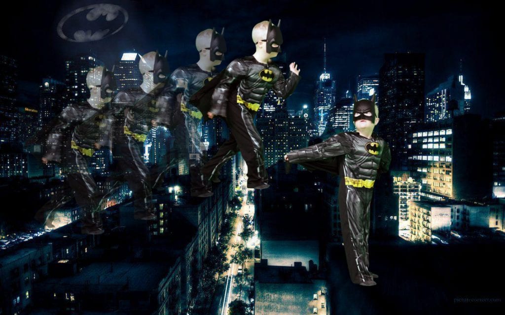 A young boy dressed in a Batman outfit stands against a backdrop of city streets. The bat signal is in the sky.