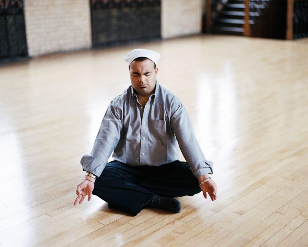 An adult with Down's syndrome sits cross-legged on the floor and is meditating
