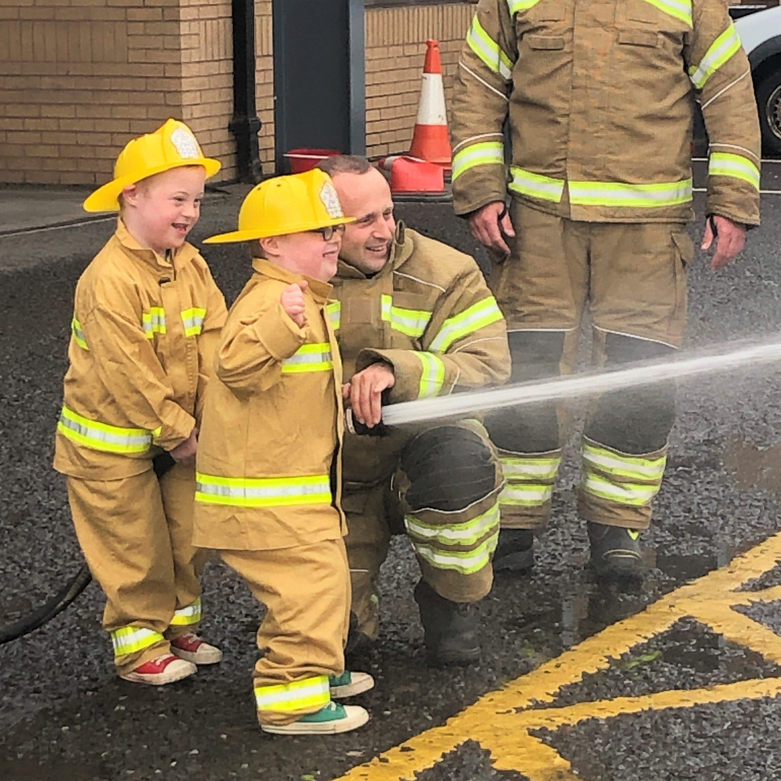 Two little boys in firemen's outfits and two firemen hold a hose on full blast