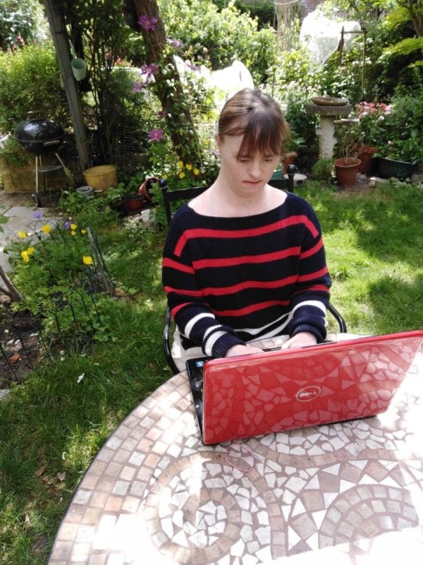 A woman sits at a table outside in a garden. She is working on a laptop. The woman has Down's syndrome.