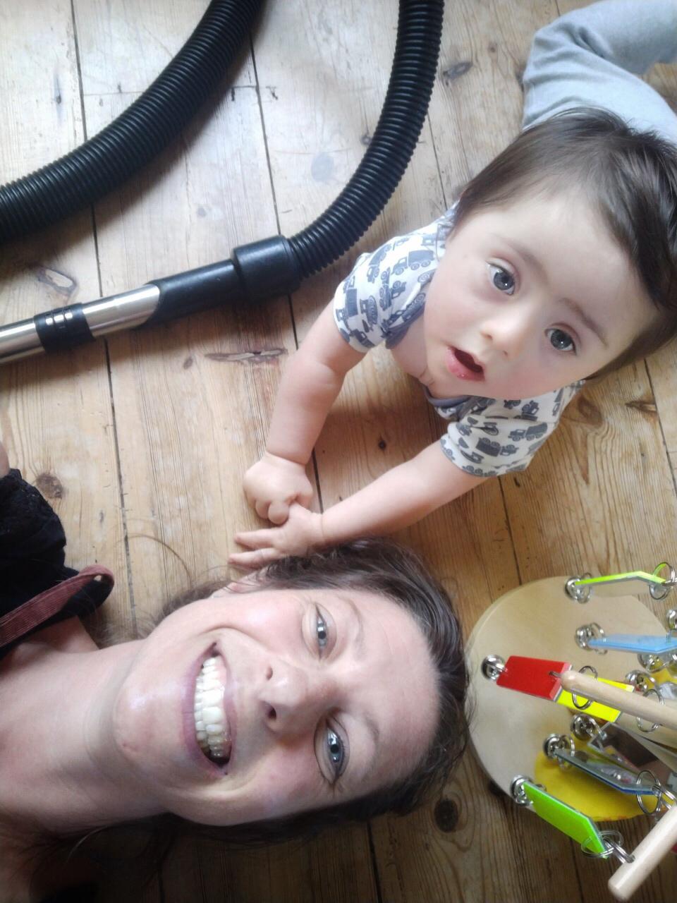 A mum lies on her back on a wooden floor. She's smiling up at the camera. Next to her is a baby who has Down's syndrome, also looking up at the camera.