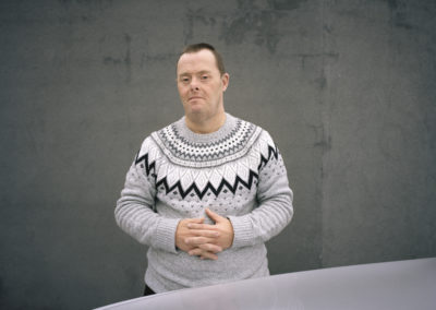 A man stares at the camera with an appraising look. He stands against a grey background and wears a grey jumper with Scandinavian designs in white and black. The man has Down's syndrome