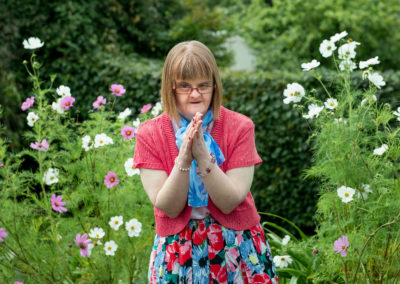 A woman wearing a flowery skirt, a pink cardigan and a blue scarf stands among clumps of cosmos flowers. She has Down's syndrome.