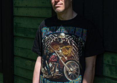The man, leaning casually against what could be a garden shed, wears jeans and a heavy metal style tshirt. He is smiling gently. He has Down's syndrome.