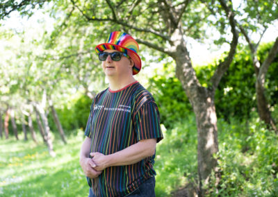Ben stands in a garden, with trees, wildflowers and a hedge in the background. He wears sunglasses, a rainbow top hat and a rainbow striped t shirt