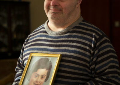 A man who has Down's syndrome stands and smiles at the camera. He is wearing a striped jumper in grey, black and dark blue. He holds a photograph of his younger self in his hands.
