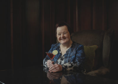 A smartly dressed woman who has Down's syndrome sits with a brightly coloured cocktail in her hand. The background is warm, dark and cosy. She smiles at the viewer.