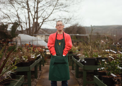 A man wearing a red sweat shirt, jeans, boots and a green apron stands in among the plants at a garden centre. His thoughtful gaze looks directly into the camera. The man has Down's syndrome.