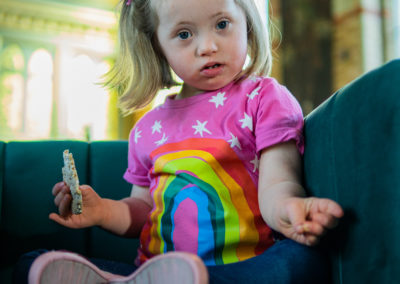 Coraline sits cross legged in an easy chair. She is looking straight at the camera. She has a rice cake in her right hand. She wears a colourful pink and rainbow tshirt and leggings and has a bow in her hair. Coraline has Down's syndrome.
