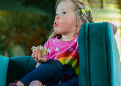 Coraline sits in an easy chair. She is gazing out of shot and has a rice cake in her right hand. Her left arm leans on the arm of the chair. She wears a colourful pink and rainbow tshirt and leggings and has a bow in her hair. Coraline has Down's syndrome.