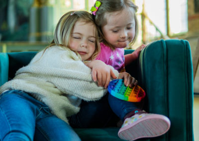 Two little girls cuddle on a sofa. One of them has Down's syndrome