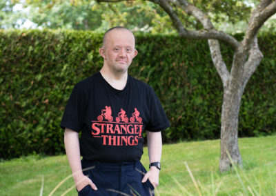 David stands in a garden, with a tree and hedge behind him. His hands rest gently in his pockets. He is wearing a Stranger Things tshirt. David has Down's syndrome.