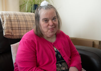A woman sits on a comfy chair and looks into the camera. She wears a floral dress, a pink cardigan and has a flower in her hair. The woman has Down's syndrome.