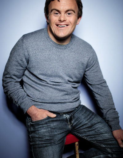 A man sits on the edge of a tall stool against a neutral background. He wears a grey sweater and jeans. He smiles into the camera, full of confidence. The man has Down's syndrome.
