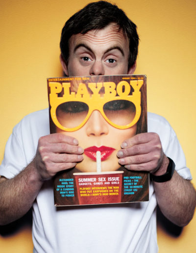 A man peers over the top of a copy of Playboy magazine, which he holds with the cover to the camera. He stands against a neutral background. His expression is quizzical and cheeky. He has Down's syndrome.