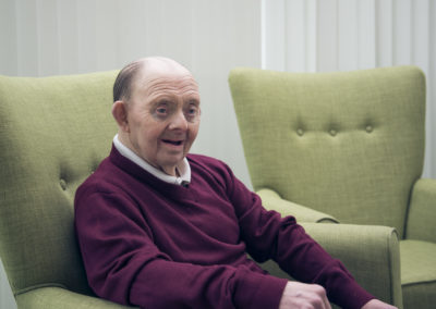 A gently smiling man sits in an easy chair. He is smartly dressed and his hands rest, one on the arm of the chair and one on his left knee. The man has Down's syndrome.
