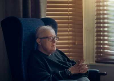 A man sits in an armchair in the soft glow of light from a partially shaded window. He sits with his hands clasped on his lap and looks thoughtful. He has Down's syndrome.