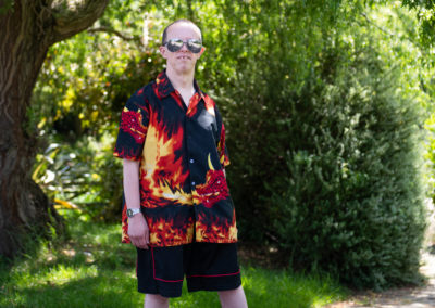 James stands in a garden. His stance is confident and relaxed. He is wearing mirrored shades and a shirt with a flaming dragon printed on it. James has Down's syndrome.