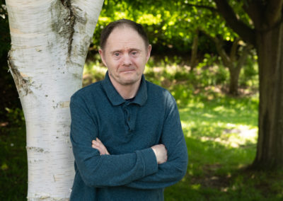 James stands in front of a silver birch tree. He looks uncertain and has his arms crossed. James has Down's syndrome.