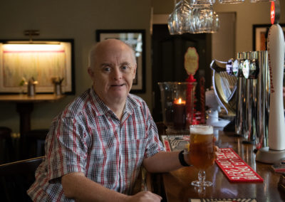 John sits at a bar with a pint of lager in his hand. He's smiling at the camera. He is wearing a cheque shirt. John has Down's syndrome.