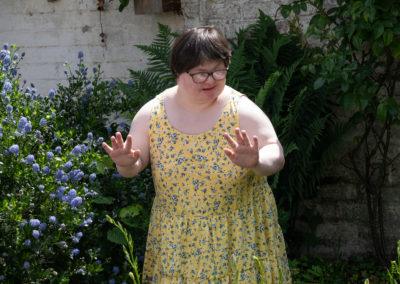 A woman in a yellow and blue flowery dress stands in the corner of a garden, surrounded by flowering shrubs and ferns. The woman has Down's syndrome.
