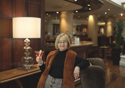A smiling woman leans against the back of an easy chair and holds a cocktail in her other hand. She stands in a hotel lounge.