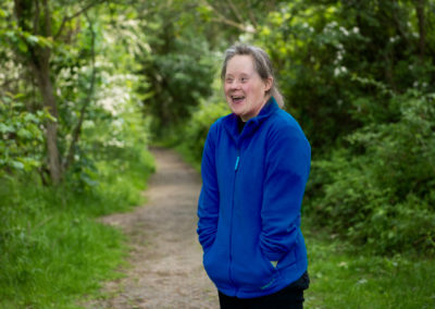 Julie stands on a wooded footpath and smiles in delight at something out of shot. She is wearing a blue fleece jacket an her hands are in her pockets. Julie has Down's syndrome.