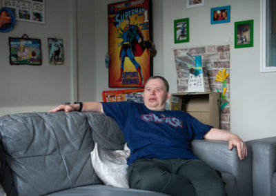 Lee sits, relaxed, on a sofa, in a room full of posters and photographs. Lee, who has Down's syndrome, wears a Grease tshirt.