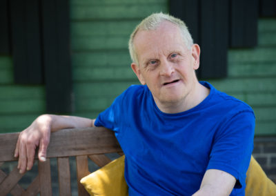 Mark sits on a garden bench, resting his right arm over the back of the bench. He wears a bright blue tshirt that brigs out the colour of his eyes that are looking directly at the camera. Mark has Down's syndrome.