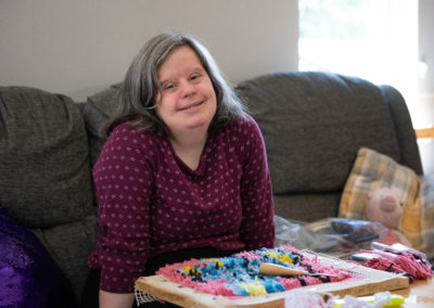 Michelle sits on a sofa and smiles gently at the camera. She has a piece of handiwork on the table in front of her. Michelle has Down's syndrome.