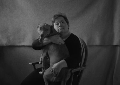 A woman sits on a wooden chair against a canvas background. She holds a dog on her lap. She is looking, curiously, directly into the camera. She has Down's syndrome.