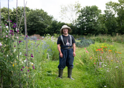 Nicholas smiles at the camera. He wears tshirt, trousers and wellington boots and a hat. He is surrounded by flowers. Nicholas has Down's syndrome.