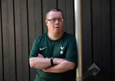 Nicky leans nonchalantly against a metal pillar. He is wearing a Tottenham shirt and has crossed arms. He is looking at something behind the photographers right shoulder. Nicky has Down's syndrome.
