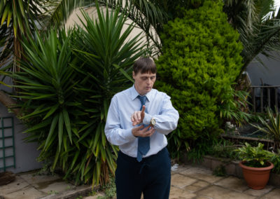 Peter stands in a garden surrounded by trees. He has a shirt and tie on and is adjusting the watch on his wrist. Peter has Down's syndrome.