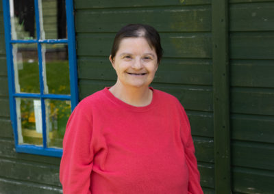 Rachel stands in front of a panelled building in a bright pink top. She smiles broadly, directly at the camera. Rachel has Down's syndrome.
