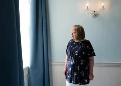 Rachel stands in the light of a large window. She looks outside with interest. She wears a flowery tunic top and white trousers. Rachel has Down's syndrome.