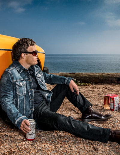 A man sits on a gravelly beach, leaning against a yellow sports car. He wears jeans, black tshirt and a denim jacket. He is wearing sunglasses. In one had he holds a can of beer...the other cans are next to him. He gazes out into the distance. The man has Down's syndrome.