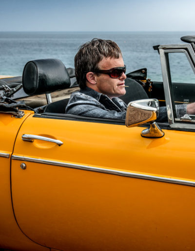 A man sits in a yellow sports car with the sea in the background. He wears a denim jacket, sunglasses and has a toothpick in his mouth. The epitome of cool. The man has Down's syndrome.