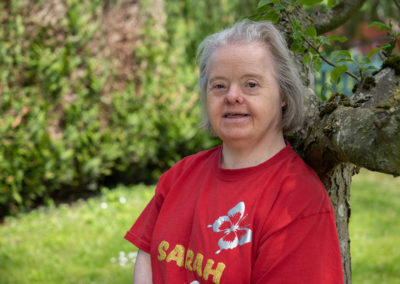 Sarah stands with her back leaning against an apple tree. She smiles warmly at the camera. She wears a read tshirt with silver butterflies and her name printed on it in golden glittery letters. Sarah has Down's syndrome.