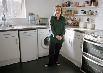 A woman smiles gently, looking at the camera. She is standing in kitchen. She holds her hands together in front of her.