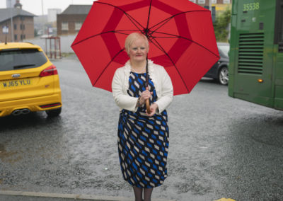 A woman holding a large, bright red open umbrella stands on the edge of the pavement. On her right is a yellow car, on her left the back end of a green bus. The woman, who has Down's syndrome, is dressed in a geometric print dress and wears a white cardigan.