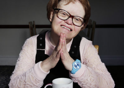 A woman who has Down's syndrome smiles into the camera. She is sitting at a table and has a mug on the table in front of her. She has her elbows on the table and her hands meet, palms together, in front of her. She is wearing glasses.