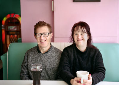 A couple sit behind a table. They both have Down's syndrome. They are both smiling into the camera. The man has a glass of coke and the woman has a mug. On the pale pink wall behind them are portraits of golden era Hollywood stars and in the background is a juke box.