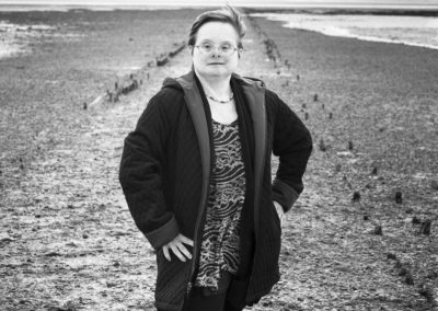 A black and white photographs of a woman who has Down's syndrome. She stands facing the camera, right hand on hip, in a coat, jeans, tunic top and boots. She stares directly into the camera. She stands on a beach or spit and there is sea in the background.